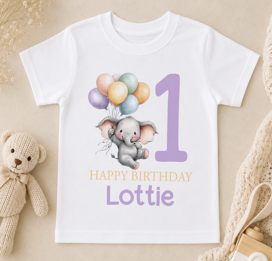 A white t-shirt with a cartoon elephant holding balloons and the number "1" printed on it, along with the text "Happy Birthday Lottie".