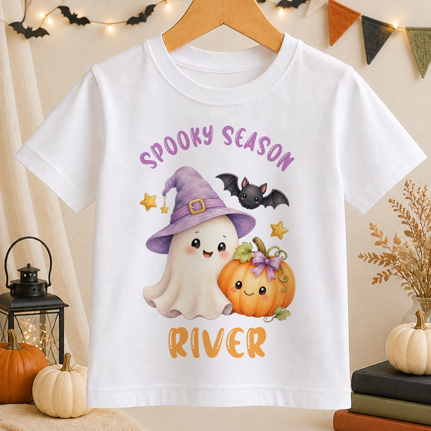 A white t-shirt with a Halloween-themed graphic design featuring a ghost, a witch, a pumpkin, and bats.