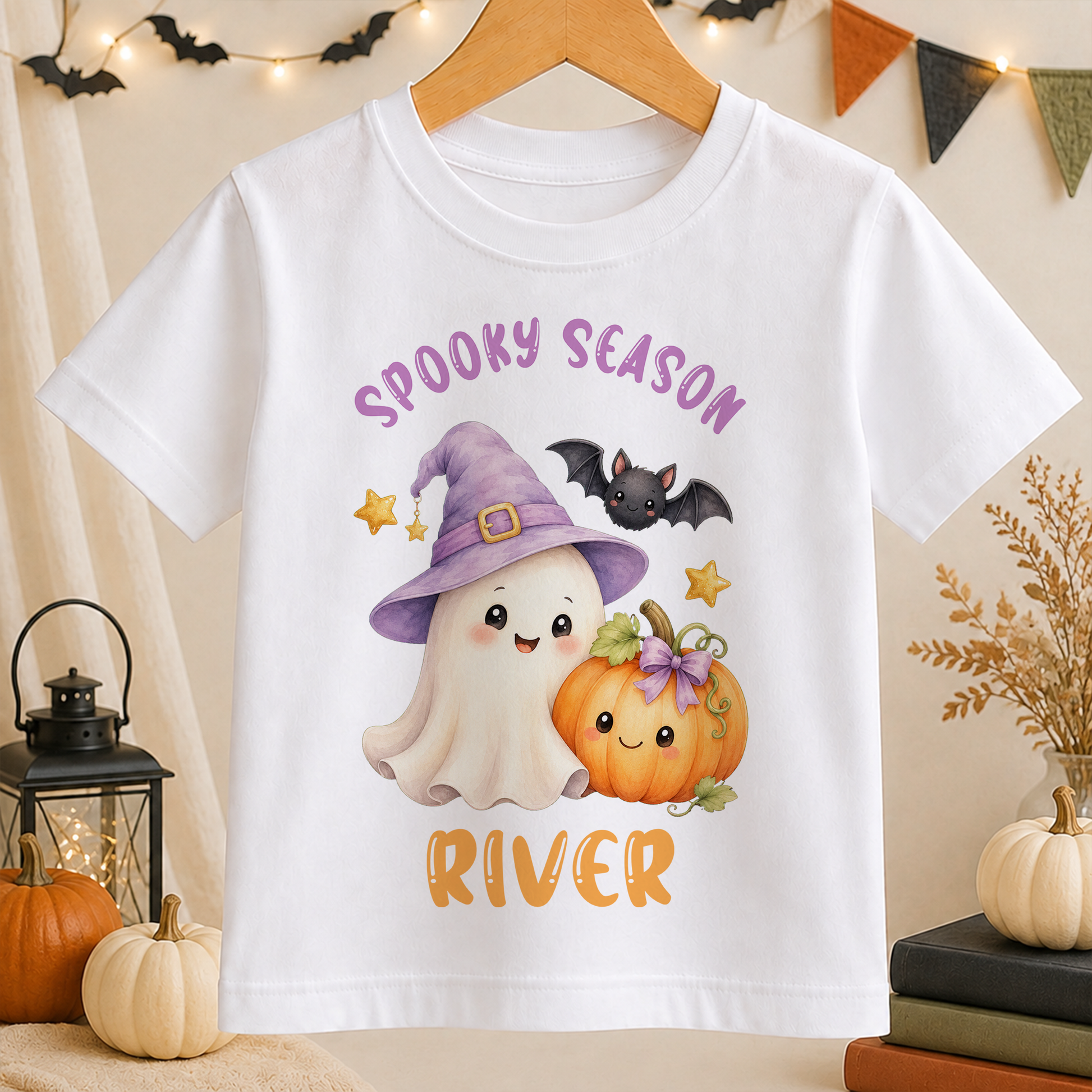 A white t-shirt with a Halloween-themed graphic design featuring a ghost, a witch, a pumpkin, and bats.