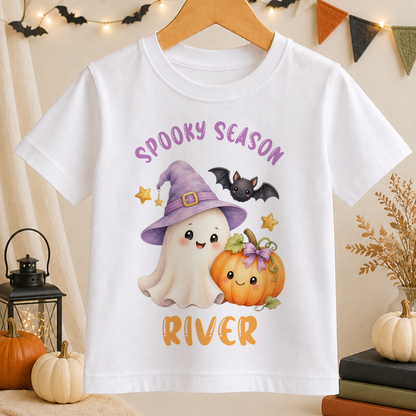A white t-shirt with a Halloween-themed graphic design featuring a ghost, a witch, a pumpkin, and bats.