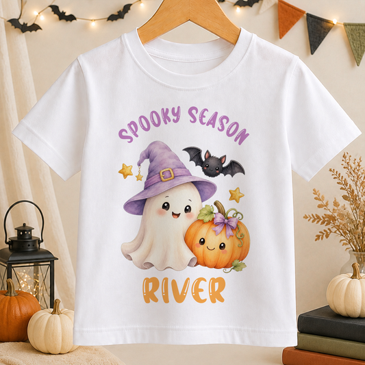 A white t-shirt with a Halloween-themed graphic design featuring a ghost, a witch, a pumpkin, and bats.