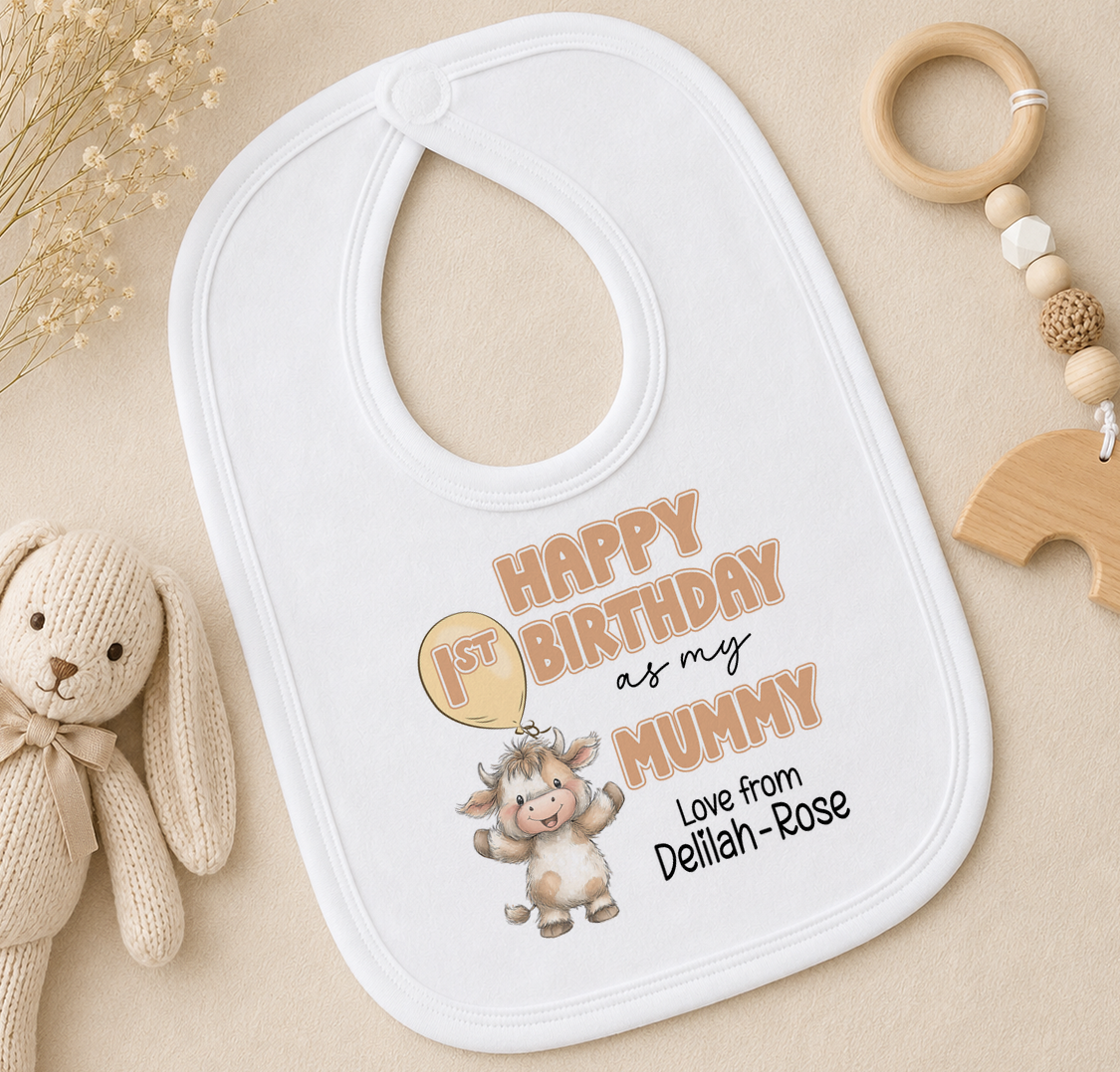 A white baby bib with a cartoon image of a cow and the text "Happy 1st Birthday as my Mummy" printed on it, along with a wooden toy and a beaded rattle.