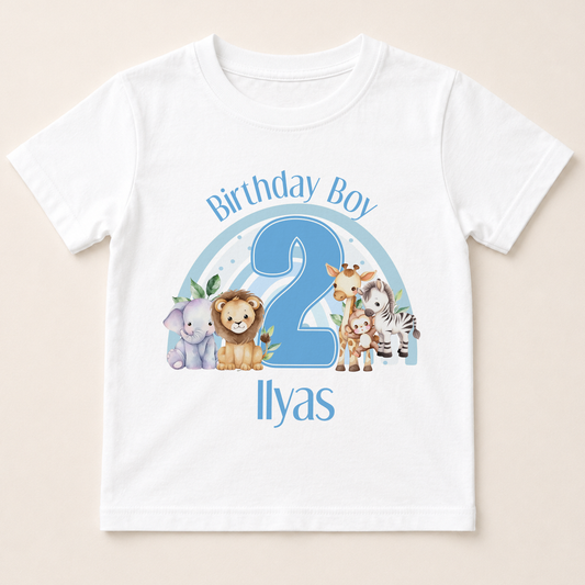 A white t-shirt with a graphic design featuring a blue number 2 surrounded by various cartoon animal characters, including a lion, zebra, giraffe, and elephant. The text "Birthday Boy 2" is written above the number, and "Ilyas" is written below the number.