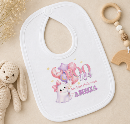A white baby bib with a personalized design featuring a ghost, balloons, and the text "My first Halloween AMELIA" is placed on a beige surface alongside a stuffed rabbit toy and a wooden bead ringer.