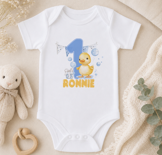 A white onesie with a cartoon duck and the number "1" printed on it, along with the text "RONNIE". The onesie is placed on a beige surface, surrounded by a stuffed animal, a wooden toy, and some greenery.