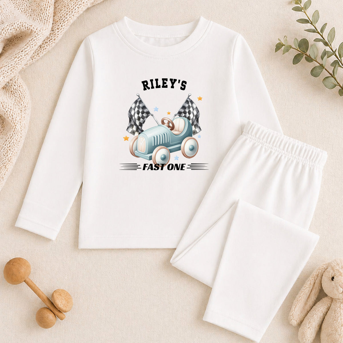 A white long-sleeved shirt with a graphic of a race car and the text "Riley's FAST ONE" printed on it, along with a pair of white pants and a stuffed animal.