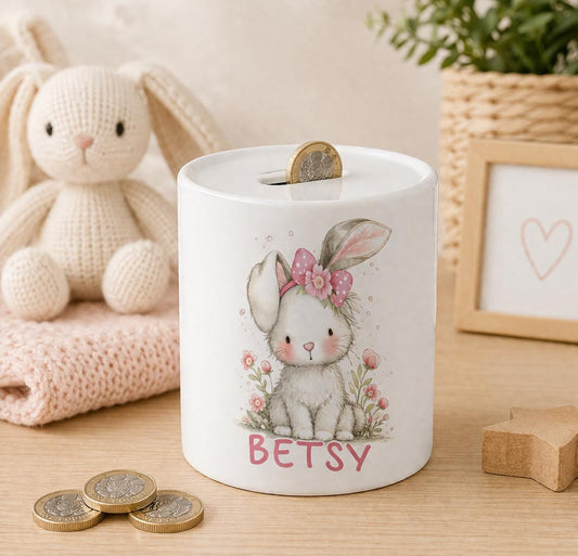 Ceramic bunny money box with a pink floral bow and personalised name in pink script, styled with a knitted bunny toy, pink blanket and coins