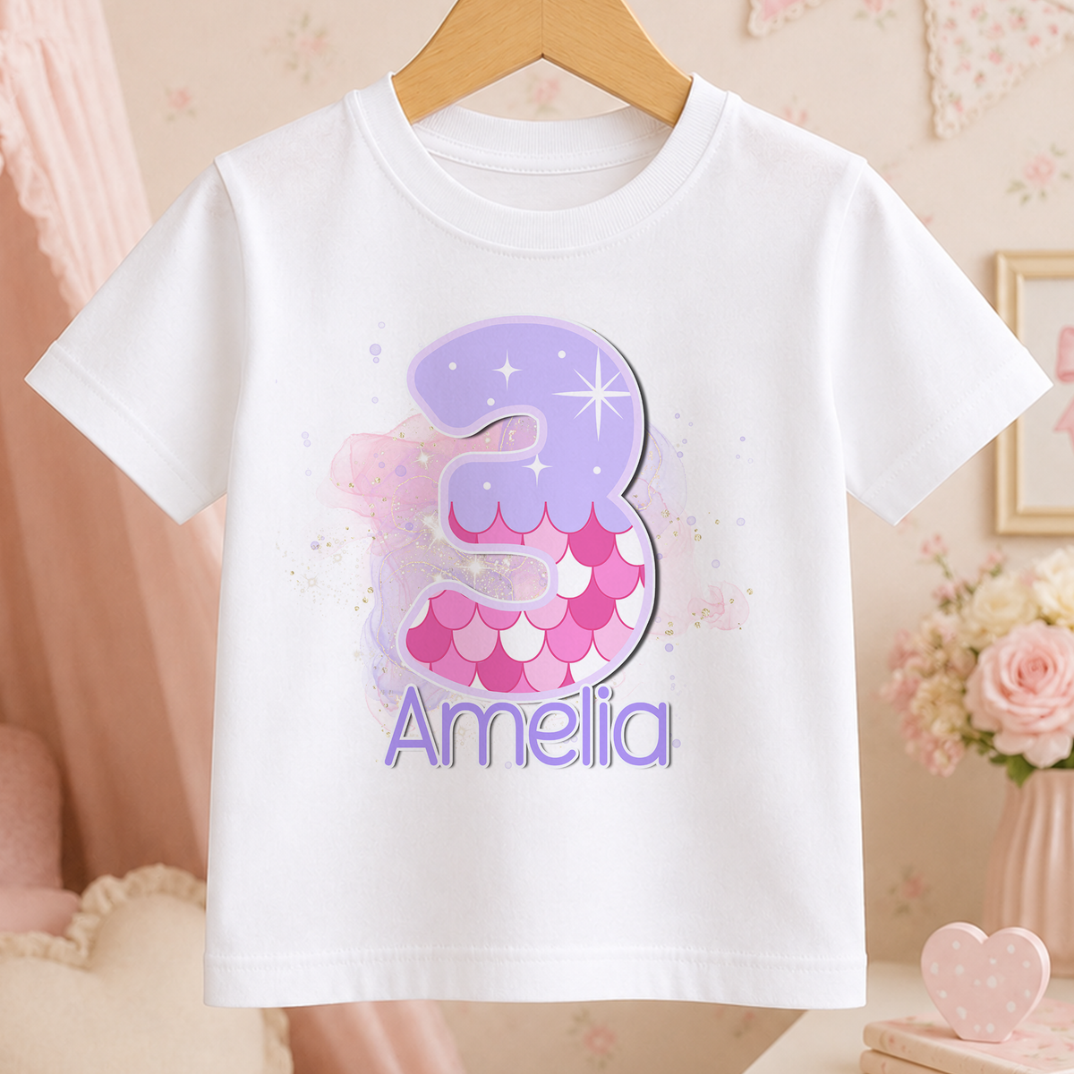 White t-shirt with a pastel mermaid birthday design, personalised number 3 and the name "Amelia", on a wooden hanger
