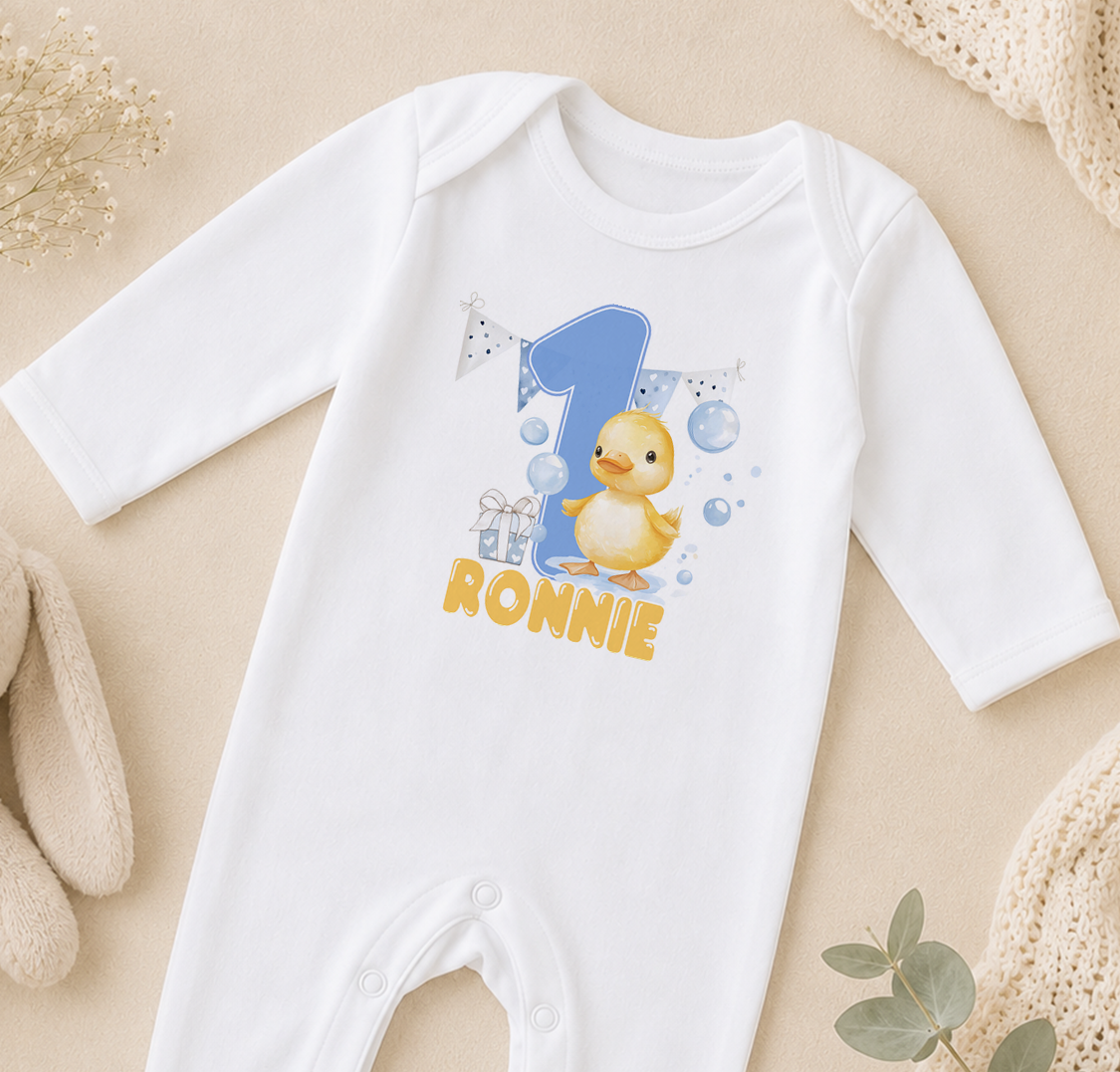 A white onesie with a cartoon duck and the number 1, along with the text "RONNIE" printed on it, is displayed on a beige surface.