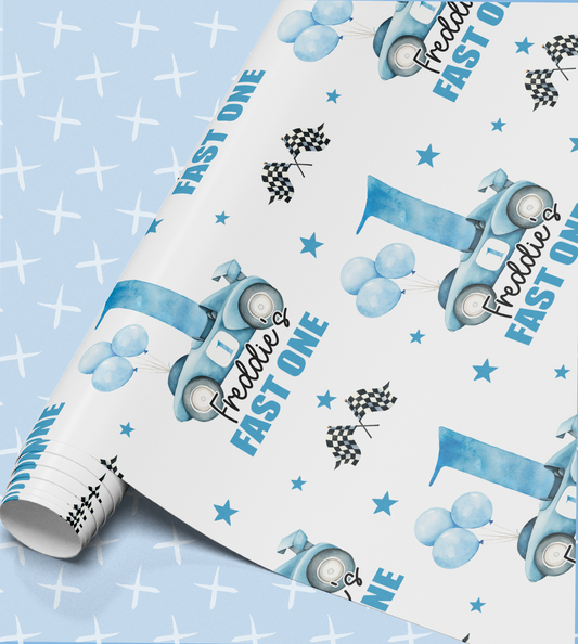 a roll of wrapping paper with a blue and white design, including a car, balloons, and stars. The text "FAST ONE" is visible, suggesting the paper is intended for a birthday celebration.