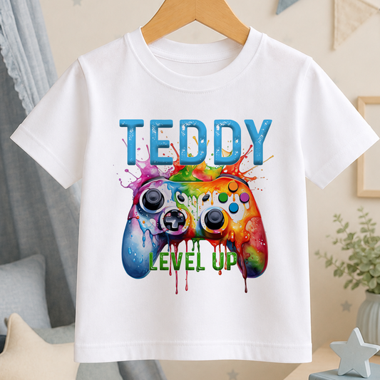 White cotton t-shirt with a colourful watercolour gaming controller print, the personalised name Teddy above it and Level Up lettering below.
