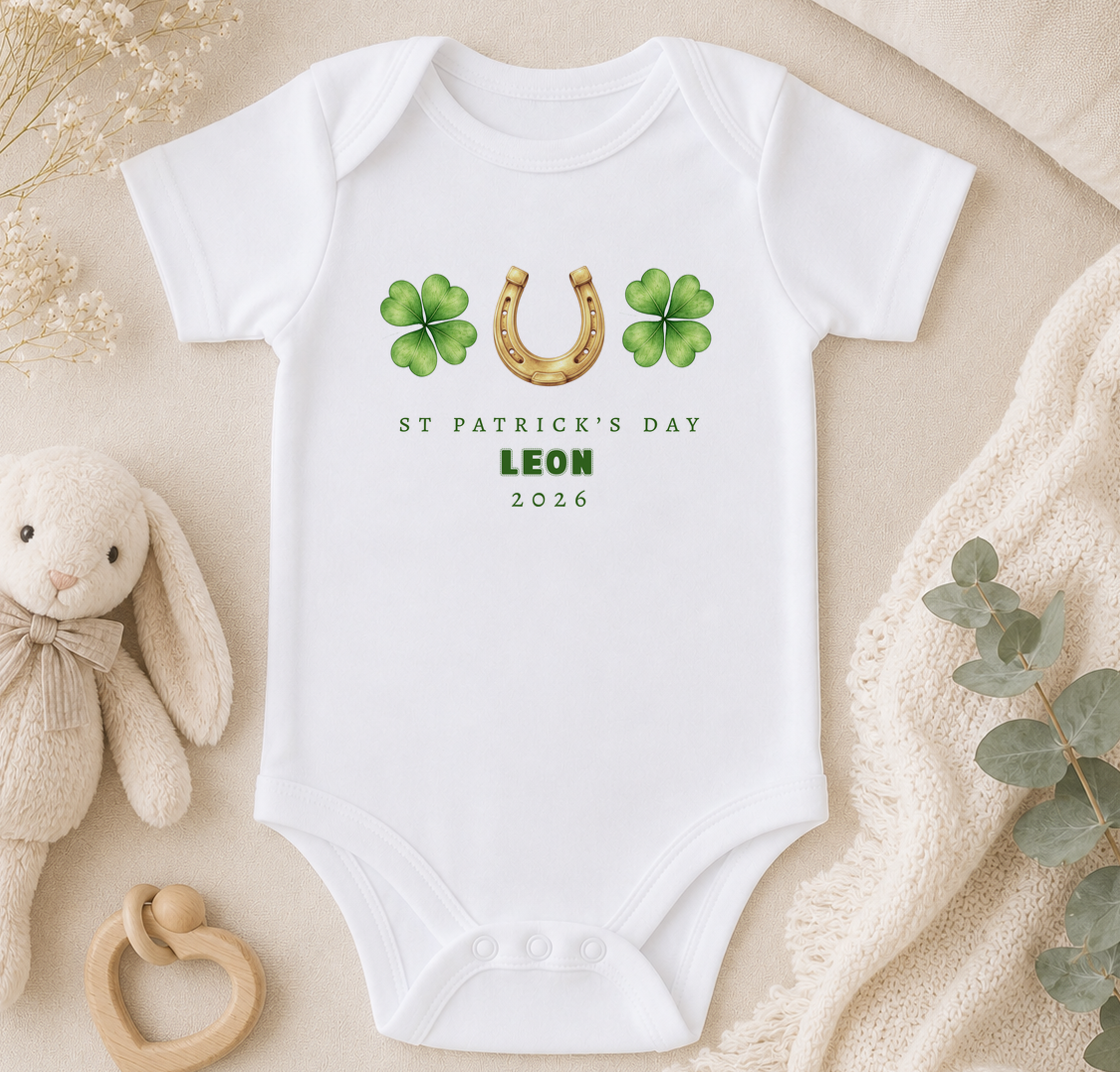 A white onesie with St. Patrick's Day and Leon printed on it, along with a gold horseshoe and four-leaf clover symbols. The onesie is placed on a beige surface, and there is a stuffed animal and some greenery nearby.