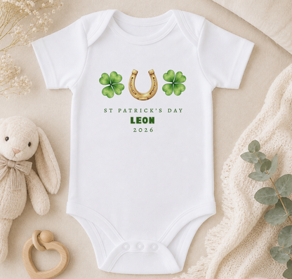 A white onesie with St. Patrick's Day and Leon printed on it, along with a gold horseshoe and four-leaf clover symbols. The onesie is placed on a beige surface, and there is a stuffed animal and some greenery nearby.