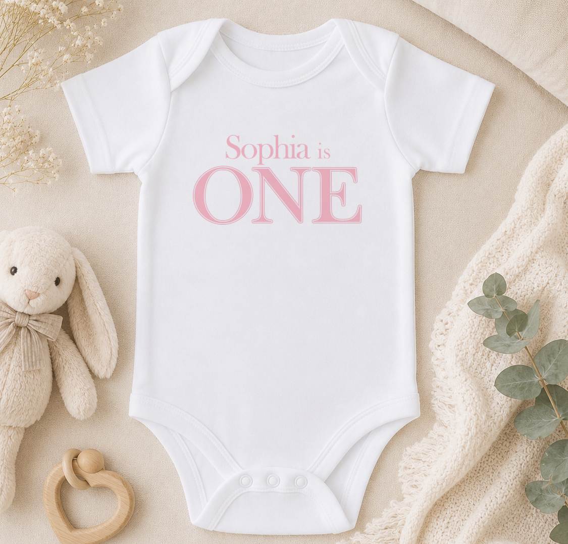 A white onesie with the text "Sophia is ONE" printed on it, along with a stuffed bunny and a wooden teething ring, all arranged on a beige surface.