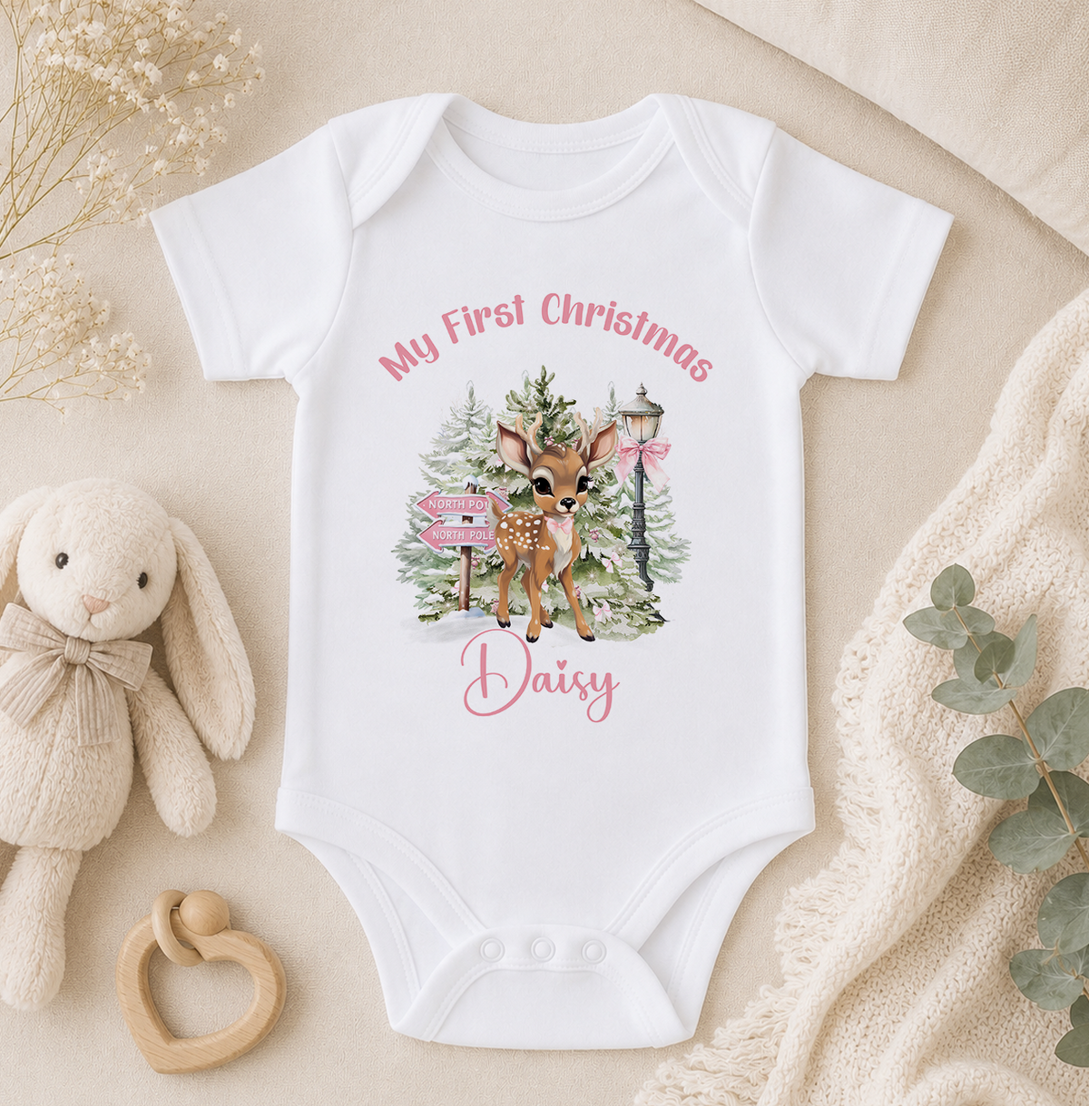 A white baby onesie with a deer graphic and the text "My First Christmas Daisy" printed on it, along with a stuffed animal and some greenery.
