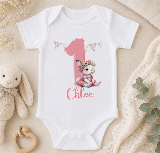 A white onesie with a pink number one and a small bunny wearing a flower crown, along with a stuffed bunny and some greenery.