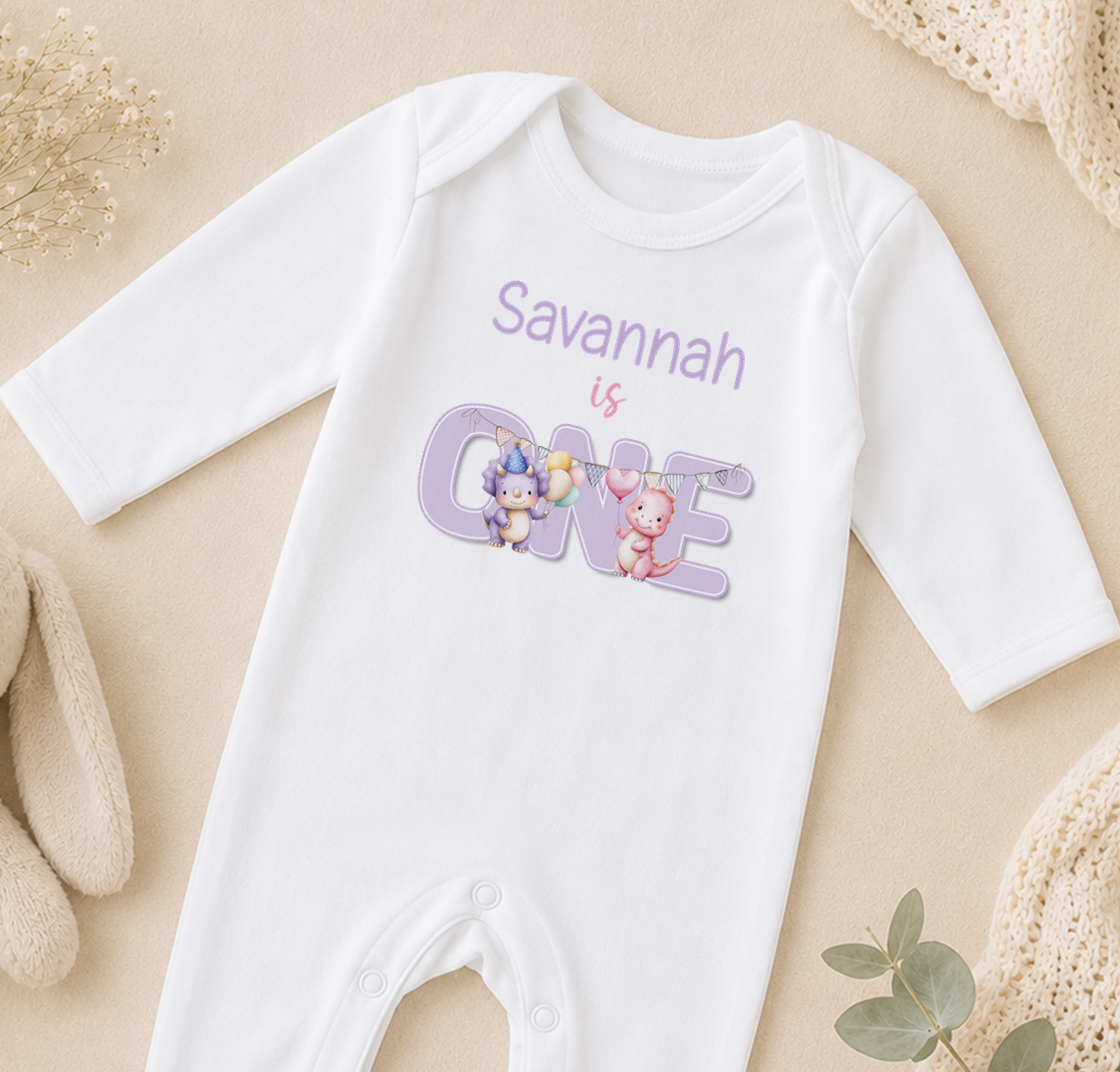 A white onesie with the text "Savannah is ONE" printed on it, along with illustrations of two cartoon animals.