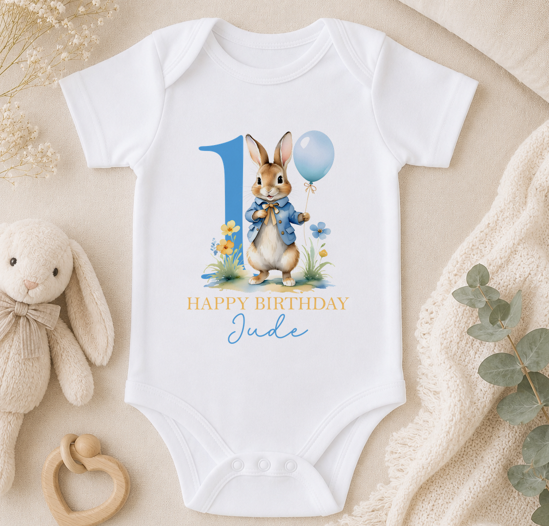 A white onesie with a bunny character and a blue balloon, along with the text "Happy Birthday Jude" printed on it, is displayed on a bed surrounded by stuffed animals and a blanket.