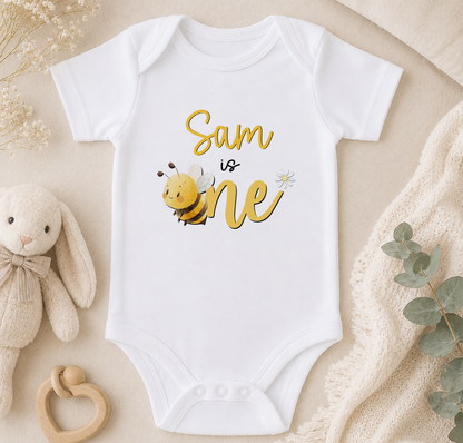 A white onesie with a bee graphic and the text "Sam is one" printed on it, along with a stuffed bunny and some greenery.