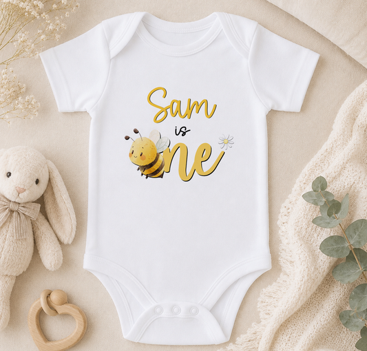 A white onesie with a bee graphic and the text "Sam is one" printed on it, along with a stuffed bunny and some greenery.