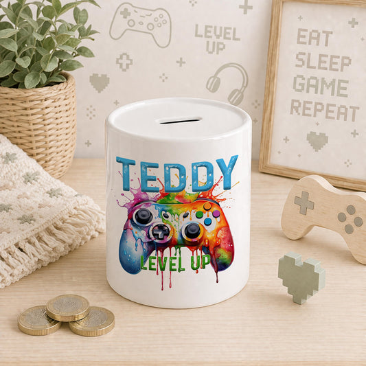 Ceramic gamer money box with a rainbow paint-splatter games controller design and Level Up text, personalised name in blue block letters, styled with a wooden controller toy and coins
