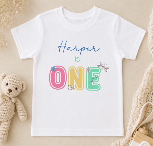 A white t-shirt with the text "Harper is ONE" printed on it, along with colorful lettering and flower designs. The t-shirt is placed on a beige surface, and there is a stuffed teddy bear and some other items in the background.