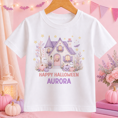 A white t-shirt with a Halloween-themed graphic design, including a house, bats, pumpkins, and a "Happy Halloween Aurora" text. The t-shirt is hanging on a wooden hanger, and there are decorative elements such as pumpkins and a garland in the background.