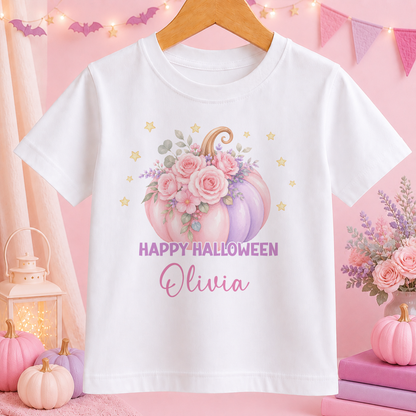 A white t-shirt with a Halloween-themed graphic of a pumpkin and flowers, along with the text "HAPPY HALLOWEEN Olivia" printed on it. The t-shirt is hanging on a wooden hanger, and there are pink pumpkins and a lantern in the background.