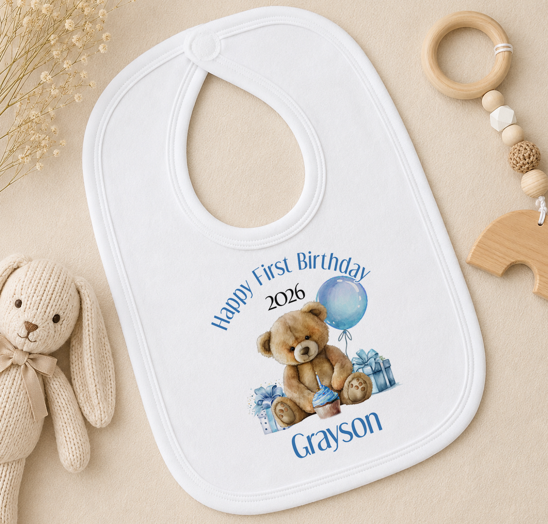 A white baby bib with a teddy bear and a blue balloon, along with a wooden toy and some dried flowers, is displayed on a beige surface.