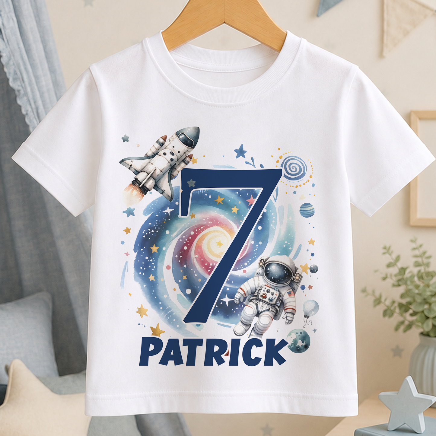 A white t-shirt with a space-themed design, featuring a rocket ship, an astronaut, and various celestial elements such as stars, planets, and a spiral galaxy.