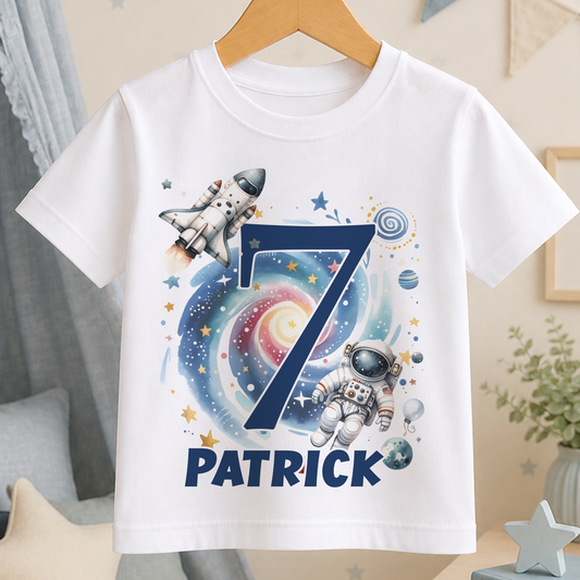 A white t-shirt with a space-themed design, featuring a rocket ship, an astronaut, and various celestial elements such as stars, planets, and a spiral galaxy.