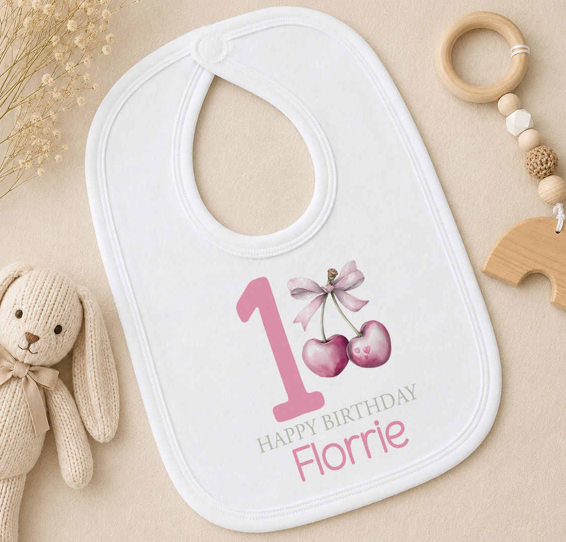 A white bib with a pink number 1 and a bow, along with a wooden toy and a beige background.
