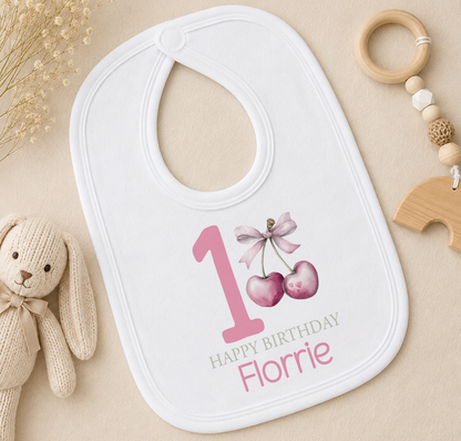 A white bib with a pink number 1 and a bow, along with a wooden toy and a beige background.