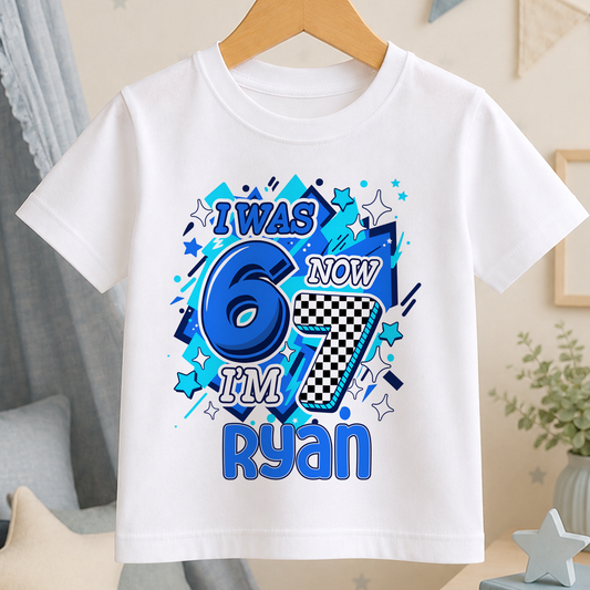 A white t-shirt with a graphic design featuring the text "I WAS 6 NOW I'M 7 RYAN" surrounded by various colorful shapes and stars.