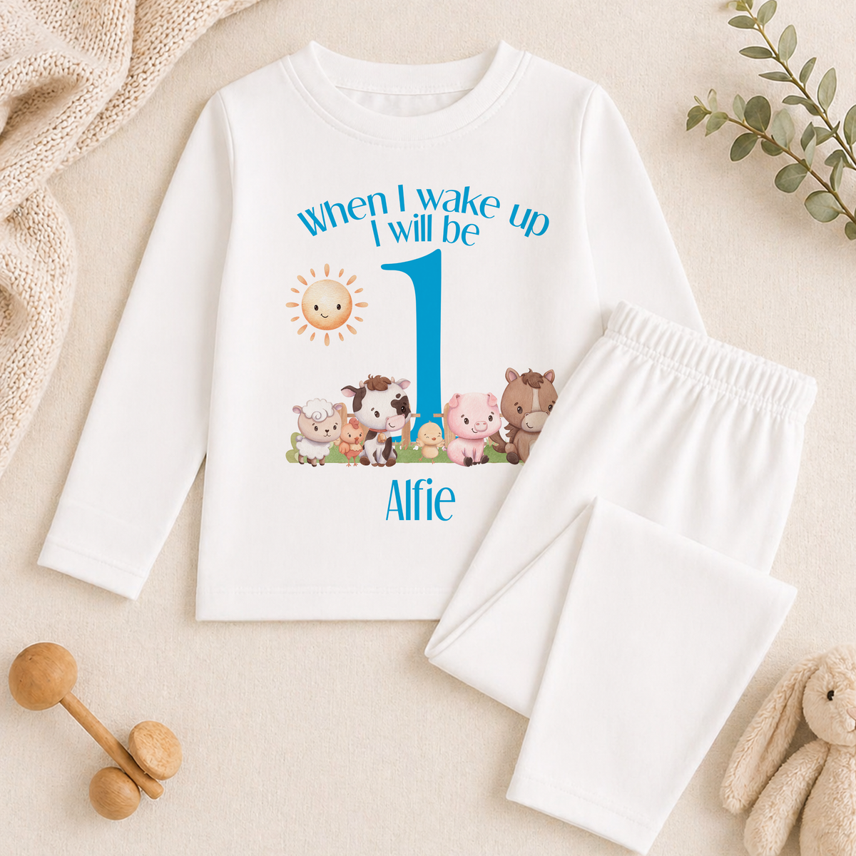 A white long-sleeved shirt with a cartoon image of farm animals and the text "When I wake up I will be 1" printed on it, along with a name tag that says "Alfie". The shirt is placed on a beige surface, and there are some toys and a plant in the background.