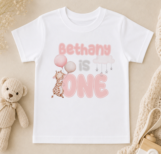 A white t-shirt with a pink and grey design that says "Bethany is one" and features a giraffe holding balloons.