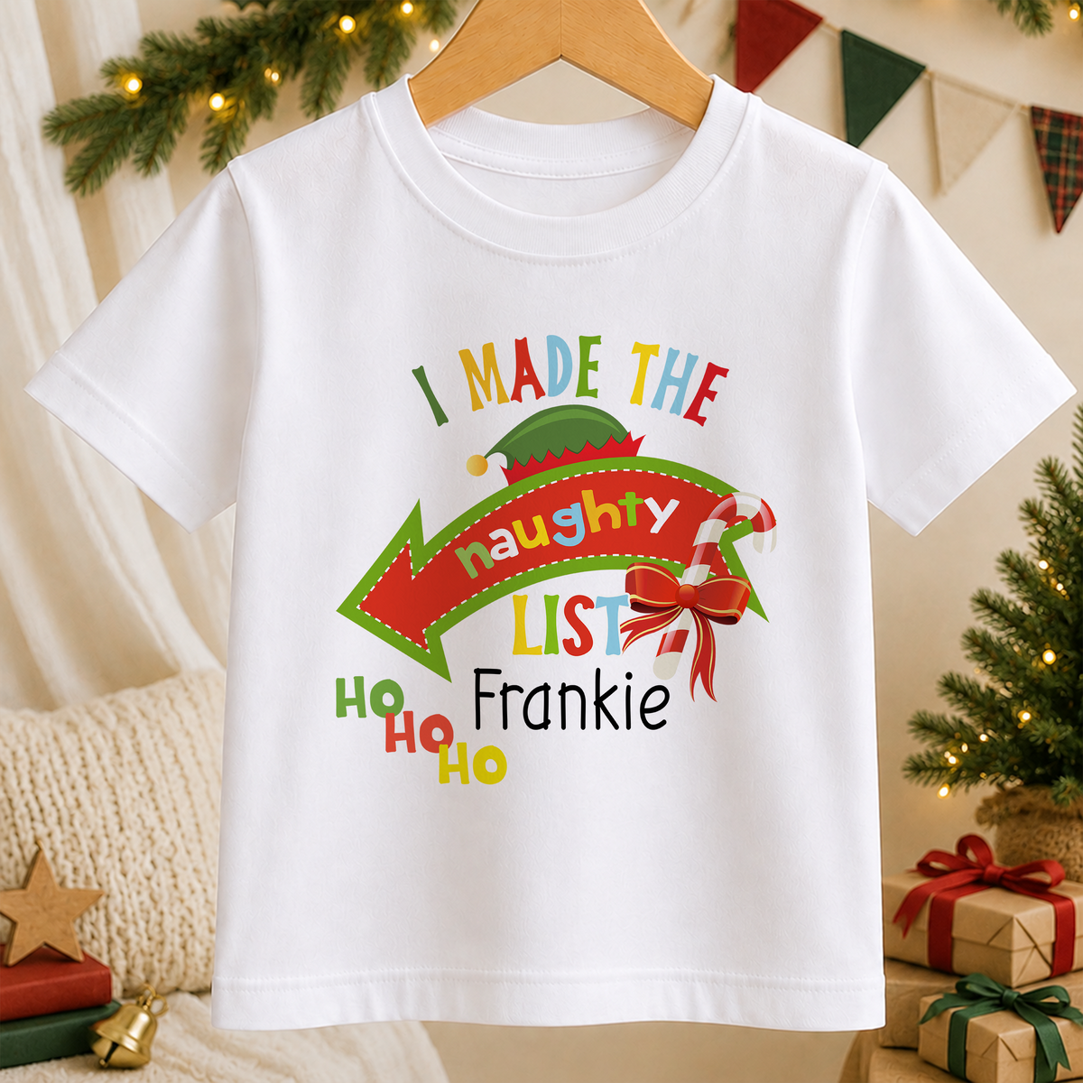 A white t-shirt with a colorful graphic design featuring a Santa hat, a red bow, and text that says "I MADE THE Naughty List" and "Frankie".