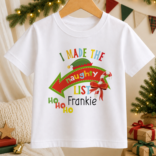 A white t-shirt with a colorful graphic design featuring a Santa hat, a red bow, and text that says "I MADE THE Naughty List" and "Frankie".