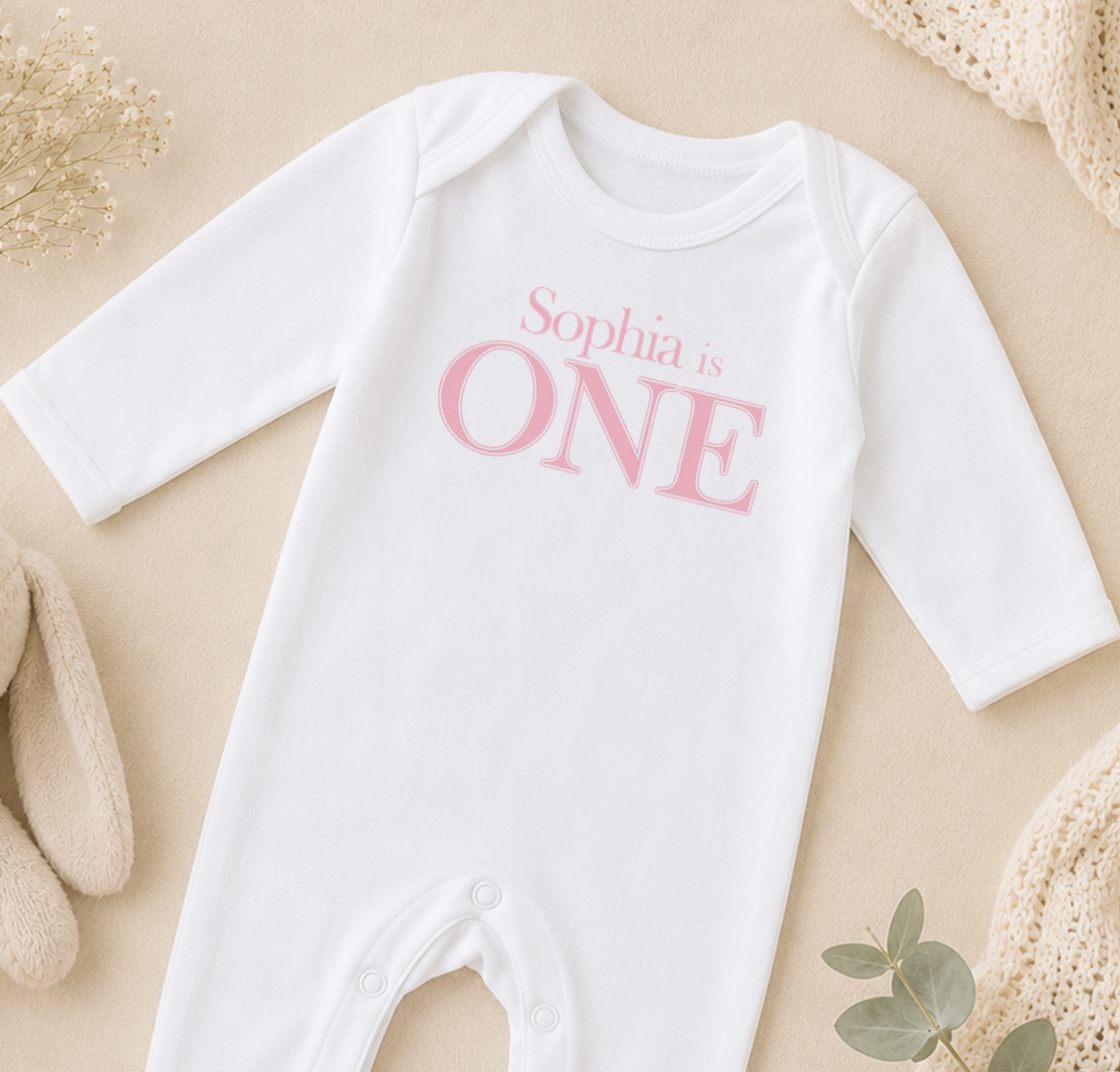 A white onesie with the text "Sophia is ONE" printed on it, laid out on a beige surface with some baby-related items such as a stuffed animal and a blanket.