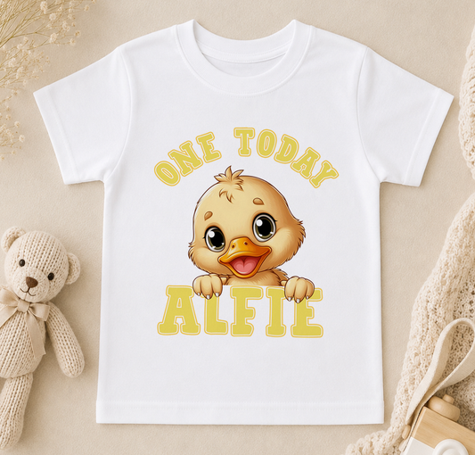 A white t-shirt with a cartoon duck and the text "ONE TODAY ALFIE" printed on it, along with a teddy bear and other baby items in the background.