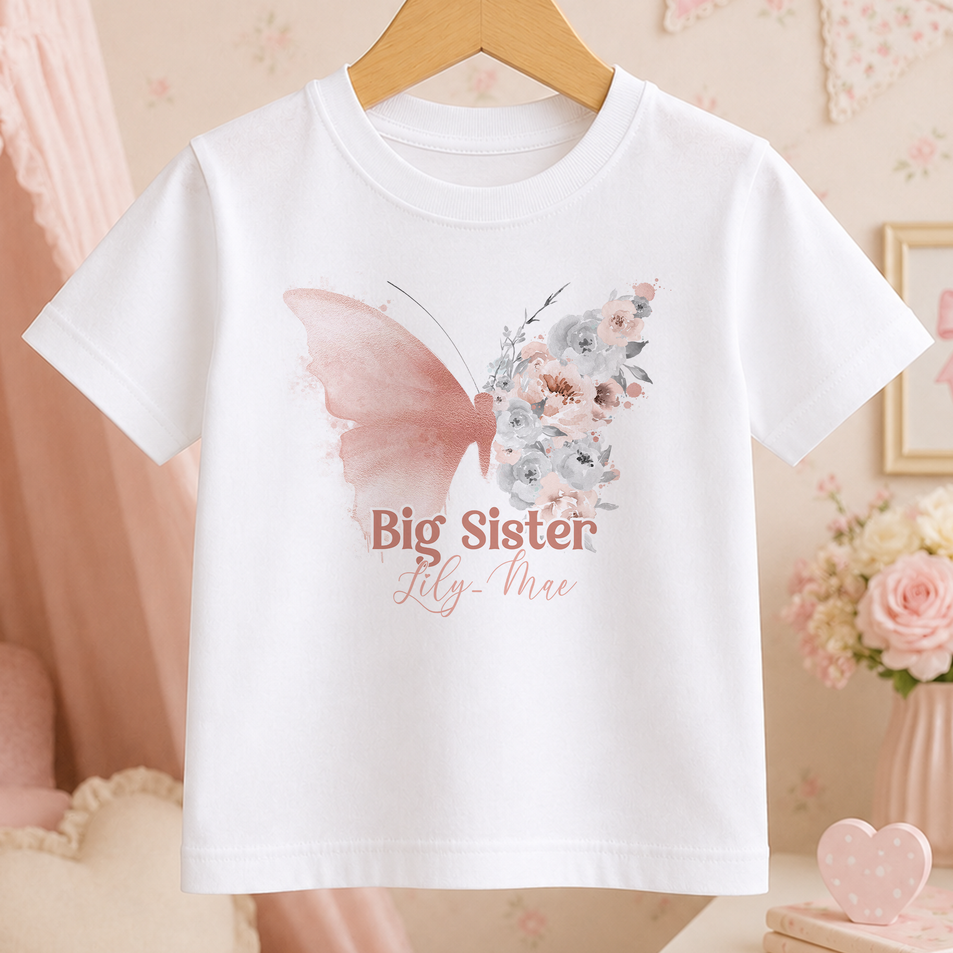 A white t-shirt with a butterfly design and the text "Big Sister Lily-Mae" printed on it, hanging on a wooden hanger.