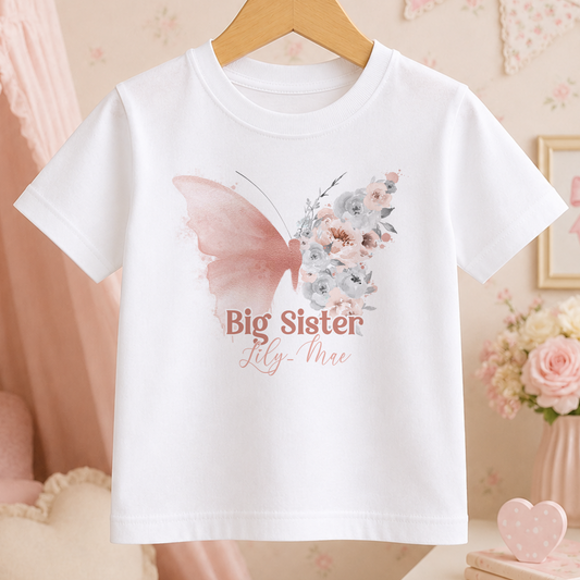 A white t-shirt with a butterfly design and the text "Big Sister Lily-Mae" printed on it, hanging on a wooden hanger.