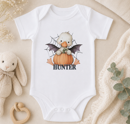 A white baby onesie with a cartoon image of a duck sitting in a pumpkin and the name "Hunter" printed on it, along with a spider web design. The onesie is placed on a beige surface, and there are some stuffed animals and a plant nearby.