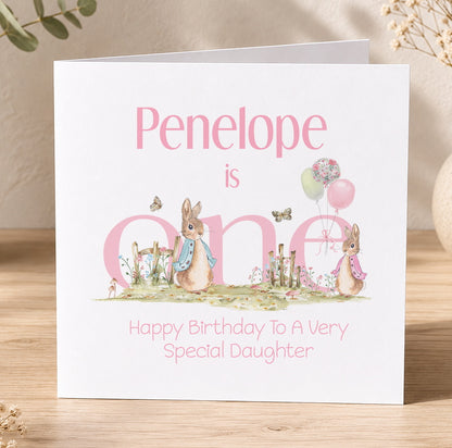Square 1st birthday card featuring a pink bunny and pastel balloons in a garden scene, personalised for a daughter's first birthday.