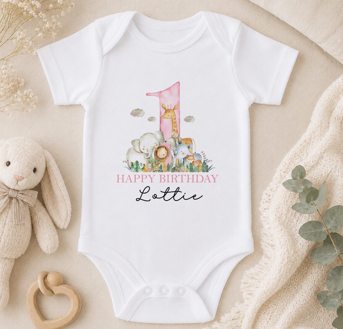 A white onesie with a pink number one and a "Happy Birthday Lottie" printed on it, along with a giraffe, elephant, and lion illustration. The onesie is placed on a beige blanket, and there is a stuffed animal and a wooden toy car nearby.
