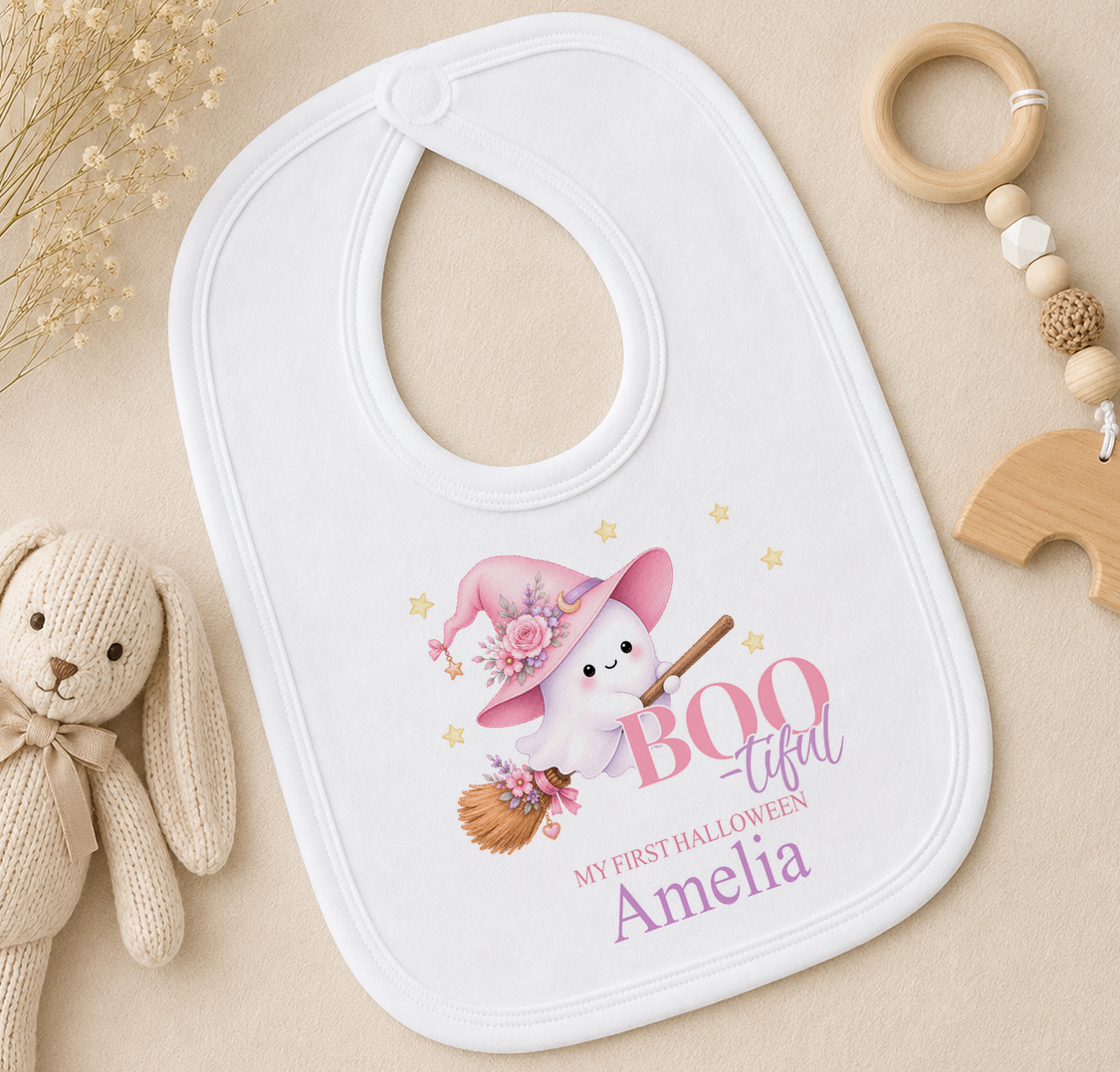A white baby bib with a cartoon ghost wearing a witch's hat and holding a broom, along with a wooden toy and a beaded rattle, all arranged on a beige surface.