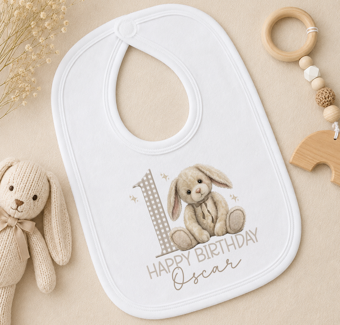 A white bib with a bunny and the number 1, along with the text "Happy Birthday Oscar", is placed on a beige surface next to a stuffed rabbit toy and a wooden bead ring.