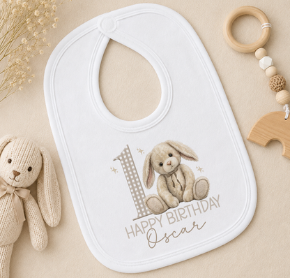 A white bib with a bunny and the number 1, along with the text "Happy Birthday Oscar", is placed on a beige surface next to a stuffed rabbit toy and a wooden bead ring.