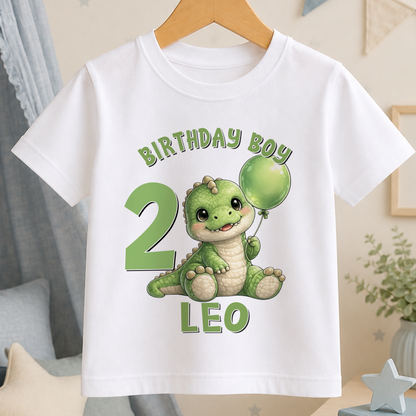 A white t-shirt with a cartoon image of a green dinosaur holding a balloon, along with the text "BIRTHDAY BOY 2 LEO".
