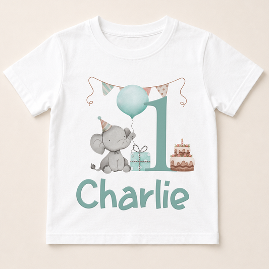 A white t-shirt with a graphic design featuring a gray elephant holding a blue balloon, a gift box, and a birthday cake. The text " Charlie" is displayed below the design.